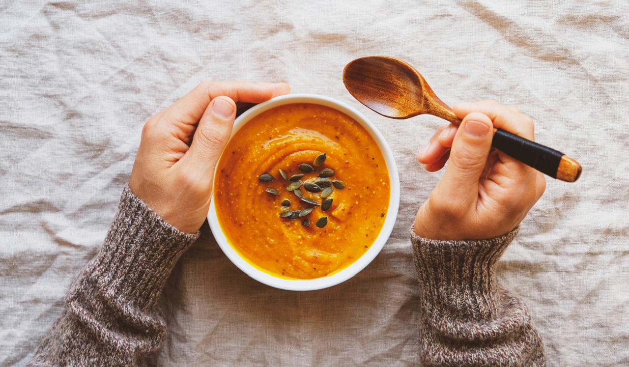 A person's hands holding a wooden spoon and a bowl of creamy orange-colored soup topped with seeds.