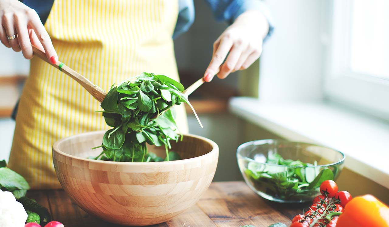 A woman wearing a yellow apron tosses a salad in a wooden bowl.