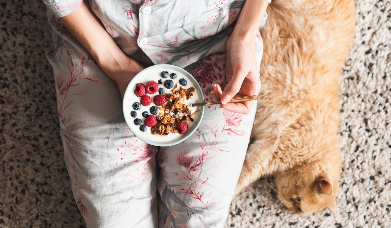 A person eats from a bowl of cereal while an orange cat lays next to them.