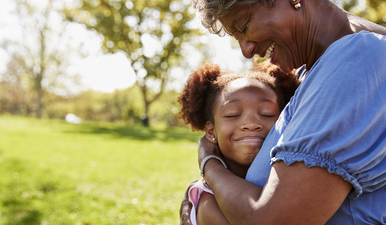 Outdoors in the sunlight, a smiling woman embraces a child lovingly.