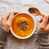 A person's hands holding a wooden spoon and a bowl of creamy orange-colored soup topped with seeds.