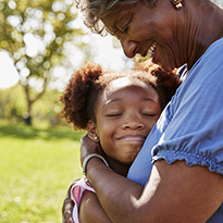 Outdoors in the sunlight, a smiling woman embraces a child lovingly.