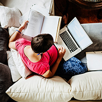 A woman on the couch with a book, notebook and laptop computer.