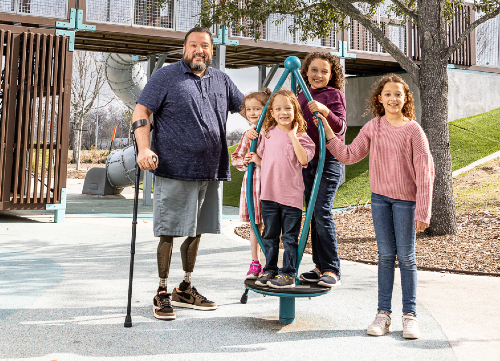 TIRR Memorial Hermann Osseointegration patient, Alex, stands with his four daughters at a play ground.