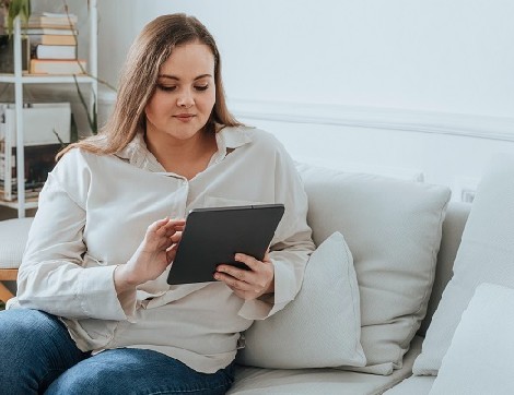 A woman sits on her couch with an electronic tablet in her hands. 